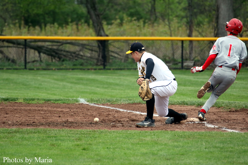 Baseball action: a catcher in a white uniform kneels at home plate with his glove down as a ground ball rolls toward him while a baserunner in gray and red slides nearby.