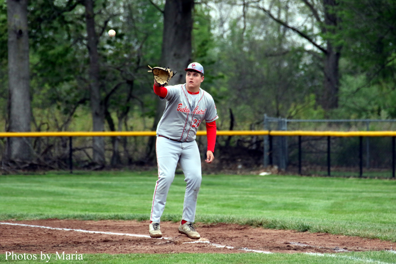 Baseball player in gray uniform with red sleeves and cap reaches with glove to catch a ball on a dirt infield.