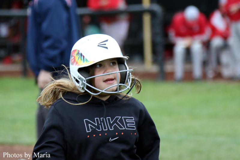 Young girl in a white batting helmet and black Nike hoodie stands on a baseball field, looking upward with a face guard in place.