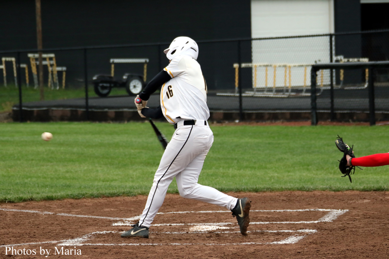 Baseball batter in white uniform swings at a pitch as the ball approaches, with catcher in red reaching nearby behind home plate.