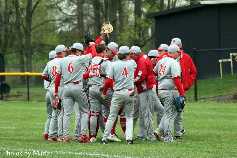 Baseball players in gray and red uniforms huddle on a green field, raising a glove in celebration.