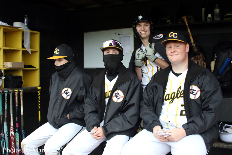 Four Eagles baseball players sit in a dugout in black jackets and white pants; two wear masks, one wears a helmet, smiling at the camera.