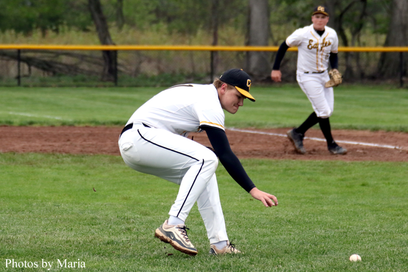 Baseball pitcher in white uniform crouches to field a ground ball on a grassy infield, with a fielder awaiting in the background.