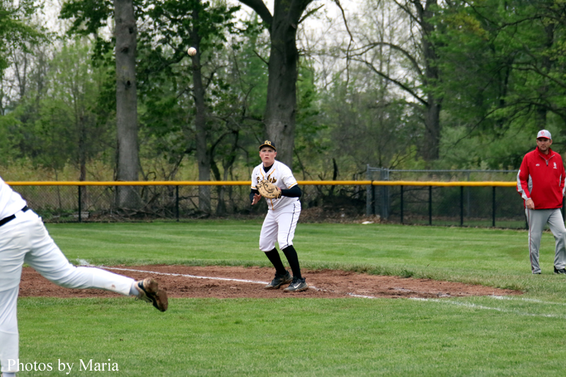 Baseball game in progress: a fielder in a white uniform with black cap and glove ready to catch a ball dropping from the air, as another player on the left runs toward the infield and a coach in red stands near the outfield fence on the right.