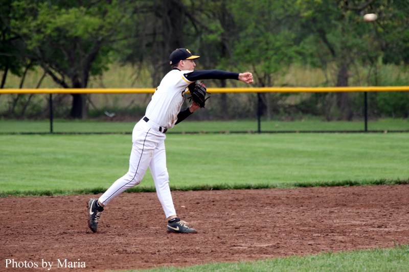 Baseball pitcher in a white uniform winds up to throw a pitch on the dirt infield with a yellow outfield fence behind him.