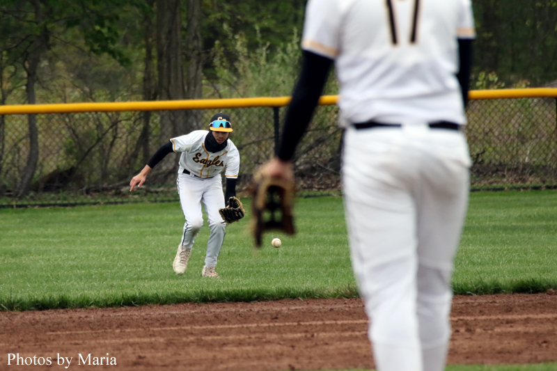 A baseball fielder in white uniform reaches for a ground ball on a grassy infield with a blurred teammate in the foreground.