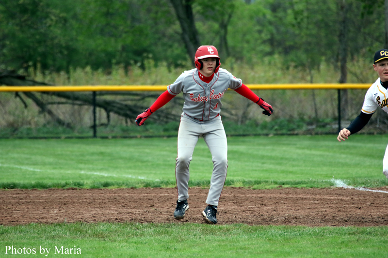 Baseball player in gray and red sliding on dirt with arms outstretched during a game, field and yellow outfield fence in the background.