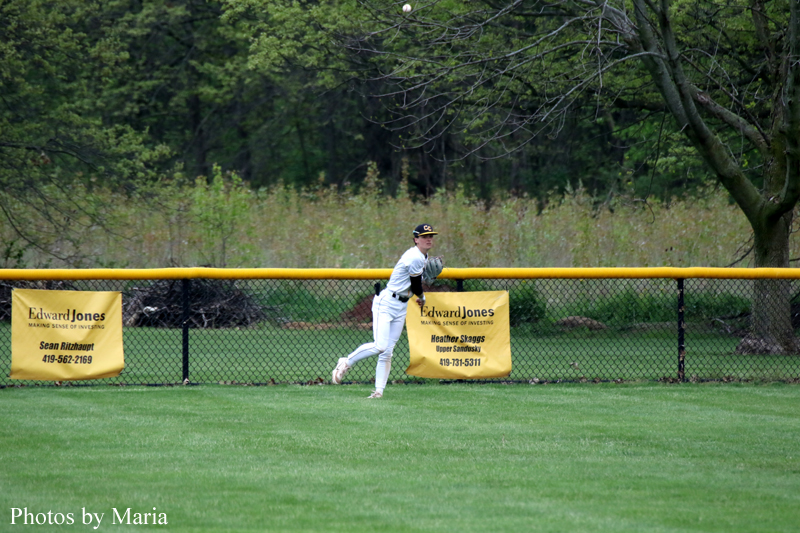 Baseball player in a white uniform runs along the outfield grass near a yellow fence with sponsor banners.