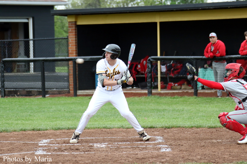 Baseball batter in white uniform standing at home plate, bat raised, ready to swing, with catcher in red gear and a dugout in the background.