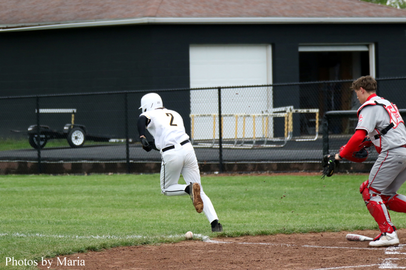 Baseball action: a white-uniformed baserunner (#2) runs toward home plate as the catcher in red gear moves in from the right. A ball sits near the dirt by the plate.