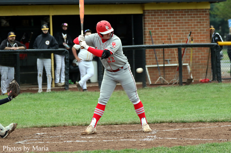 Baseball batter in gray and red swings at a pitch at home plate while teammates watch from the dugout in the background.