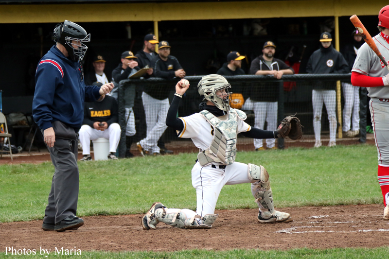 Baseball catcher in white gear kneels on dirt with fist raised in celebration as umpire looks on and teammates watch from the dugout behind.