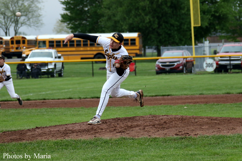 Baseball pitcher in white uniform on the mound delivering a pitch during a game, with yellow school buses and parked cars in the background.