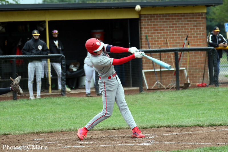 Baseball batter in gray and red swings a light-blue bat at a pitch on a dirt infield, with the dugout and teammates in the background.