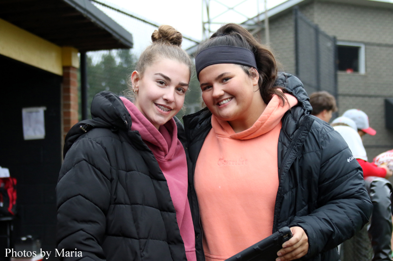Two smiling young women pose together in jackets at an outdoor event, leaning close for a photo.