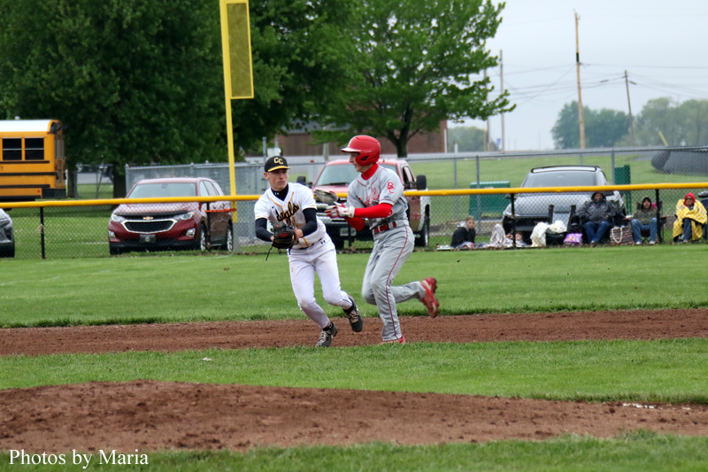 Two baseball players contest a play on the infield: a white-uniform fielder reaching for the ball as a gray-uniform runner with a red helmet slides by, with spectators and a fence in the background.