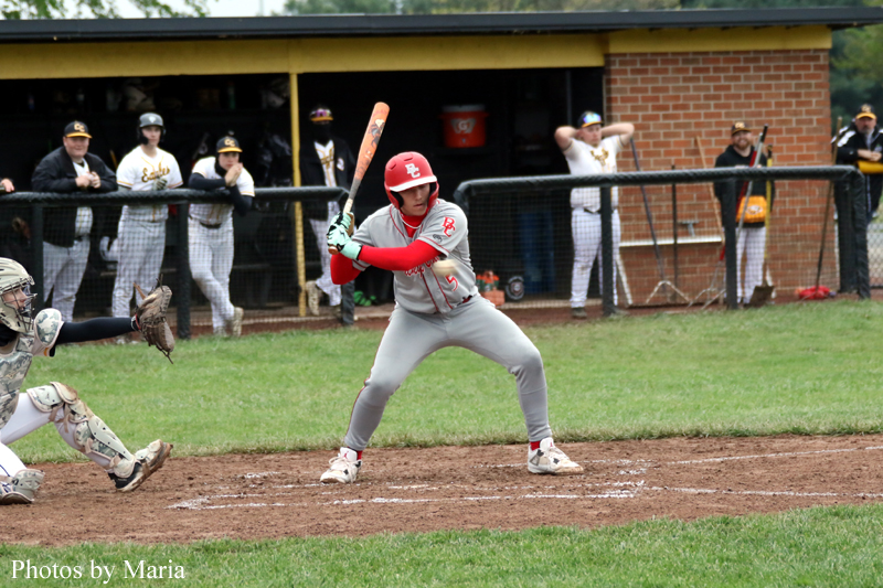 Batter at home plate in gray and red uniform, helmeted and ready to swing, with catcher and infielders behind him.