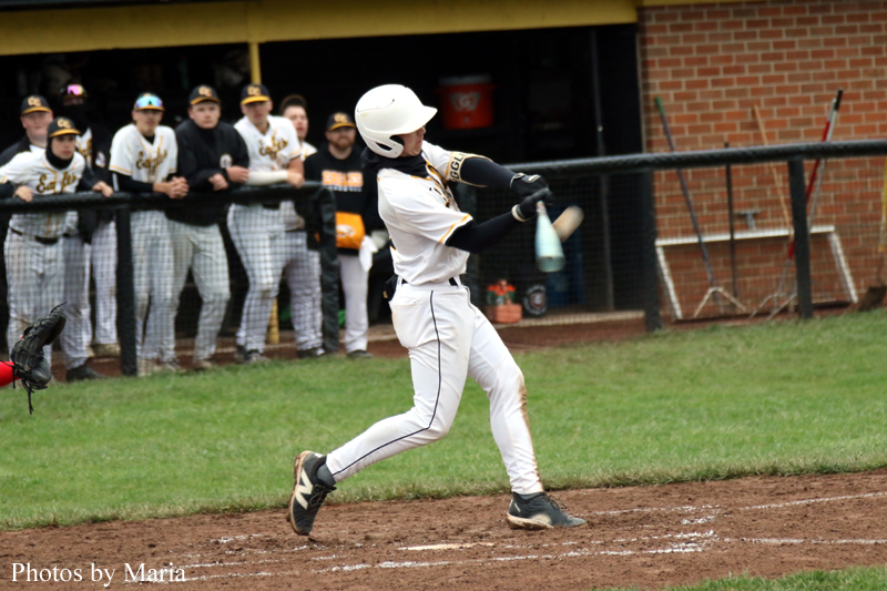 Baseball batter in white uniform swings at a pitch as teammates watch from the dugout behind him.