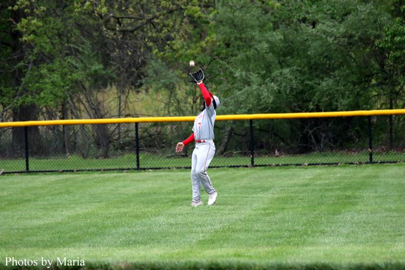 Baseball outfielder in gray and red catches a high pop foul near the yellow fence line with trees in the background.
