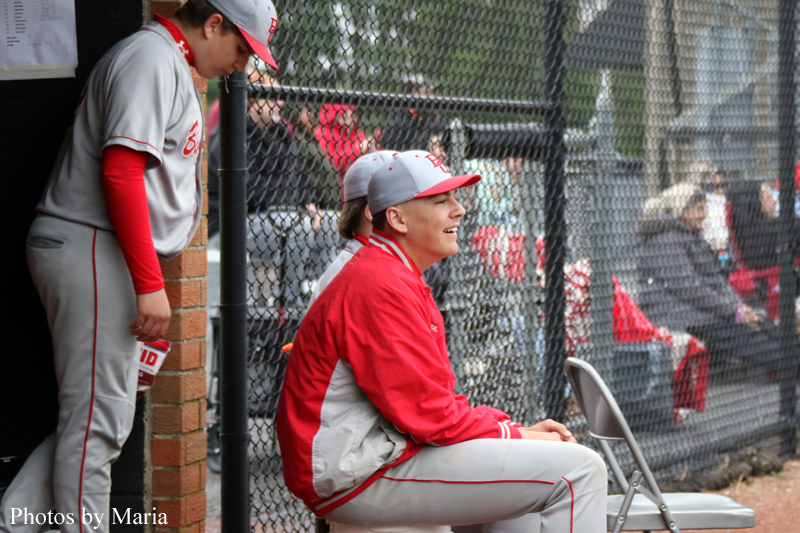 Young baseball player in red jacket sits on a folding chair by a chain-link fence, smiling, while a teammate in gray and red stands nearby by the dugout wall.