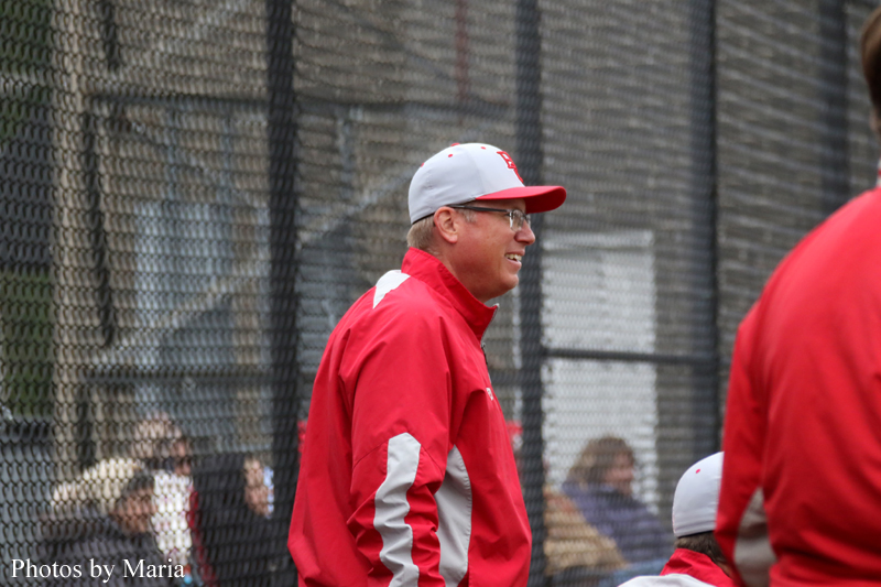 Baseball coach in a red jacket and white cap smiles while standing near a chain-link fence at a practice field.