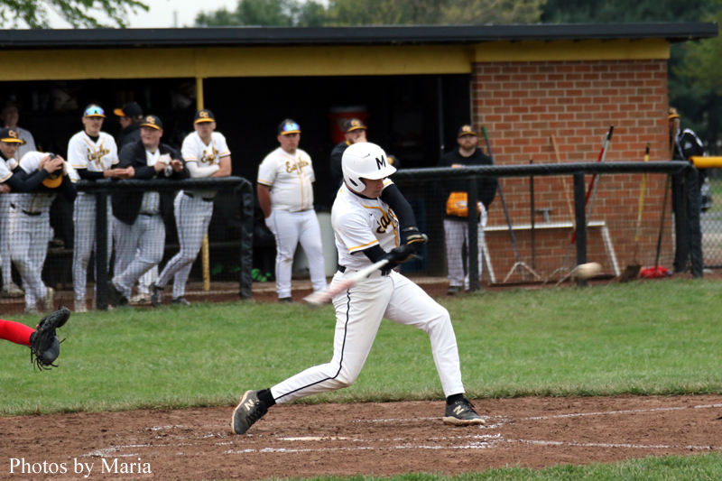 Baseball player in a white and yellow uniform swings at a pitch near home plate as teammates watch from the dugout behind a fence.