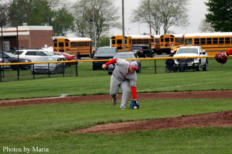 Baseball fielder in gray and red reaches for a ground ball, diving on the grass with dirt path nearby; school buses and parked cars in the background.