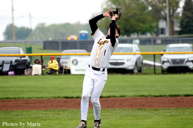 Baseball player in white-and-gold uniform reaching up with a glove to catch a high fly ball on a grassy field.