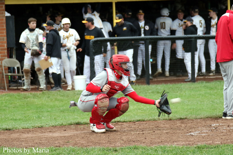 Youth baseball catcher in red gear crouches to field a ground ball near home plate, with teammates in the dugout behind him.