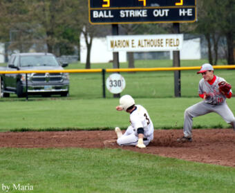 Baseball game moment: runner slides into a base as a fielder approaches on a dirt infield, with a scoreboard and Marion E. Althouse Field sign in the background.