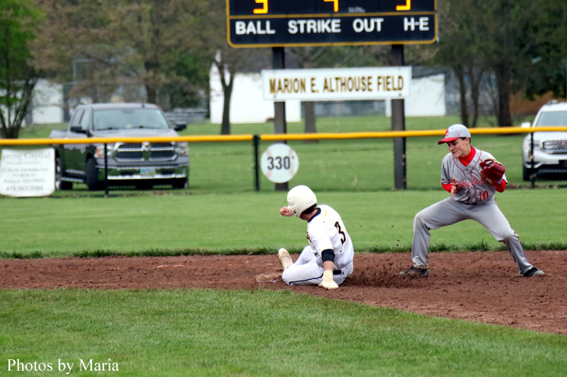 Baseball game moment: runner slides into a base as a fielder approaches on a dirt infield, with a scoreboard and Marion E. Althouse Field sign in the background.