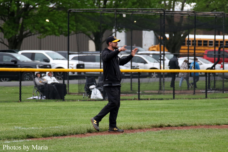 Baseball coach in black signals with hands on the infield dirt, standing on the grass near the foul line as a fence and spectators watch behind him.