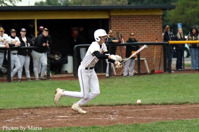 Baseball batter in white uniform swings at a pitch near home plate as teammates and spectators watch from the dugout and fence.