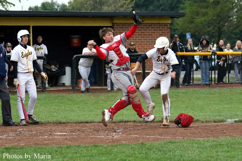 Catcher in red gear leaps over a sliding baserunner at home plate; helmet sits on dirt as a fielder in white watches.