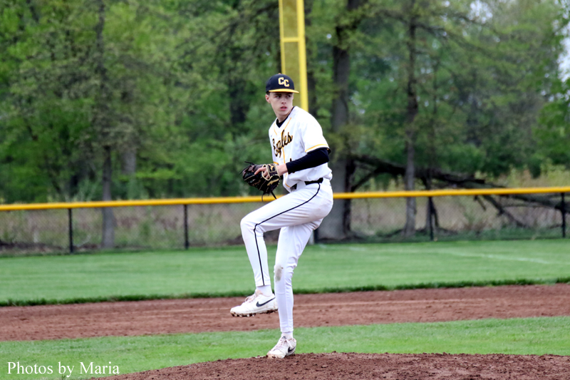 Baseball pitcher on the mound in white and gold uniform, glove up, mid-pitch with a green field and fence behind.