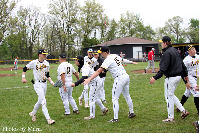 Baseball team in white and black-and-gold uniforms high-fiving on a grassy field after a game, with a small clubhouse in the background and trees around.