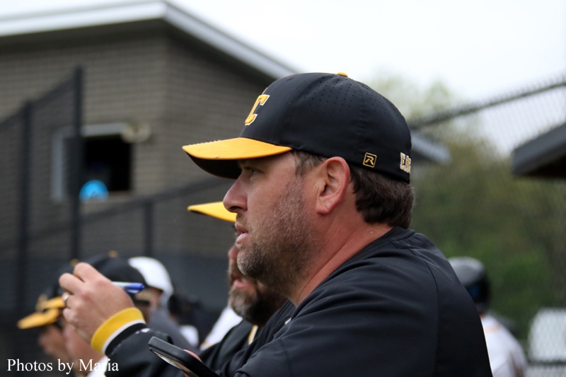 Profile of a bearded man in a black cap with a yellow brim watching a game from the dugout, holding a phone in his hand.