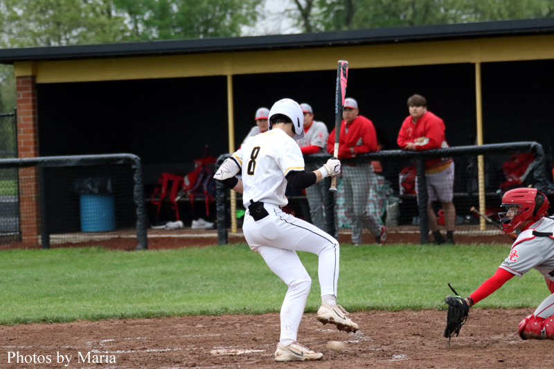 Baseball batter in a white uniform mid-swing at home plate, with a red-gear catcher crouched nearby and a dugout in the background.