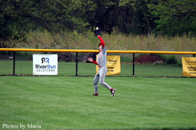 Baseball player in gray uniform leaps with glove raised to catch a fly ball on a grass field with a yellow outfield fence and sponsor banners behind.