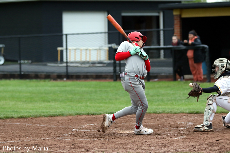 Young baseball batter in a gray uniform and red helmet swings a red bat at a pitch, with a catcher ready behind him near home plate.