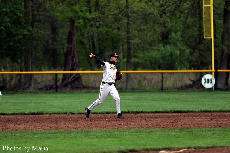 Baseball pitcher in white uniform delivering a pitch on the mound with a green outfield and fence behind him.
