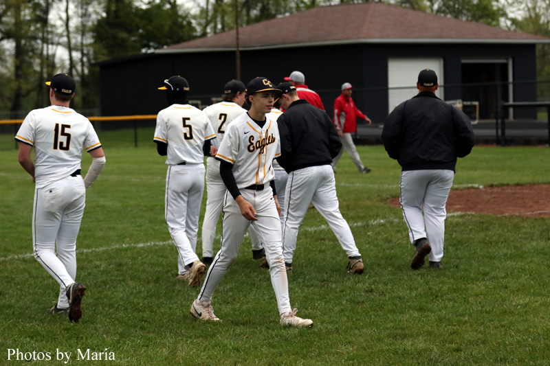 Baseball players in white and gold uniforms walk across a grassy field, Eagles on their jerseys, with a dark dugout in the background.