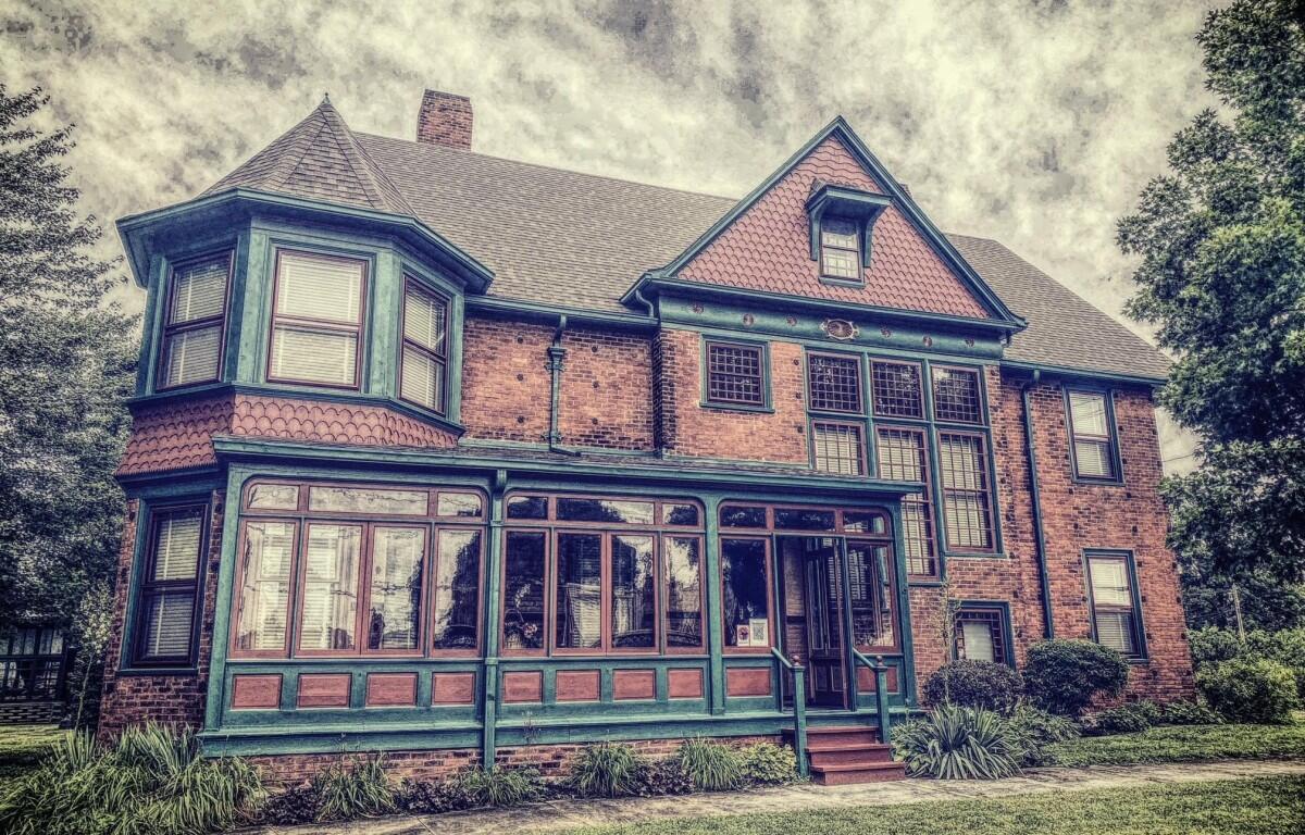 Brick two-story house with teal trim and a long, glass-enclosed porch; vintage architectural details and a manicured lawn.