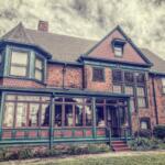 Brick two-story house with teal trim and a long, glass-enclosed porch; vintage architectural details and a manicured lawn.