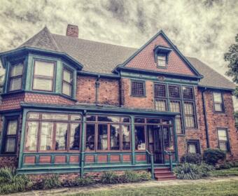 Brick two-story house with teal trim and a long, glass-enclosed porch; vintage architectural details and a manicured lawn.