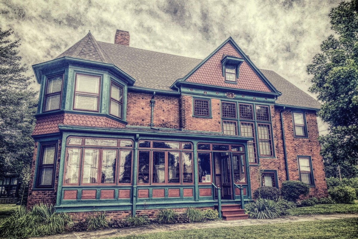 Brick two-story house with teal trim and a long, glass-enclosed porch; vintage architectural details and a manicured lawn.