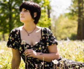 Person with short dark hair wearing a black floral dress sits in a sunlit meadow, holding a white dandelion.