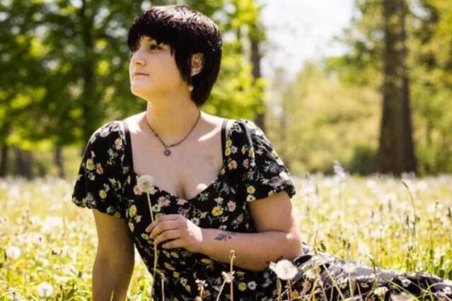 Person with short dark hair wearing a black floral dress sits in a sunlit meadow, holding a white dandelion.