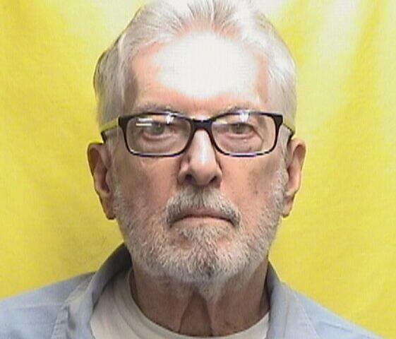 Portrait of an older man with white hair and a short beard, wearing glasses, against a bright yellow background.