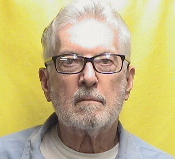 Portrait of an older man with white hair and a short beard, wearing glasses, against a bright yellow background.